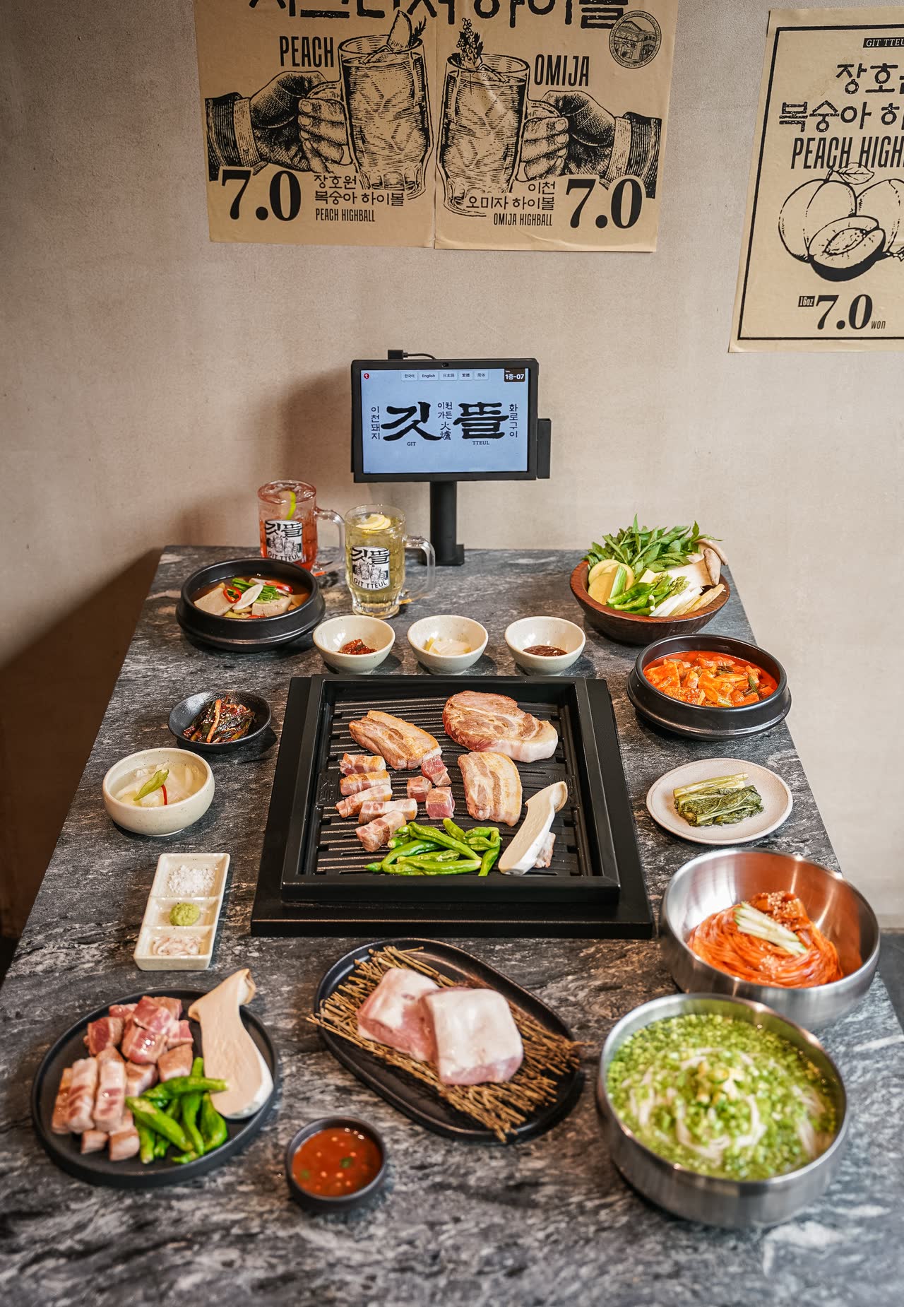 위에서 내려다본 한 상. 가운데 화로가 놓인 테이블에 생고기 접시와 김치, 찌개, 나물 반찬, 하이볼 한 잔이 깃뜰 로고가 들어간 깔개 위로 차려져 있다