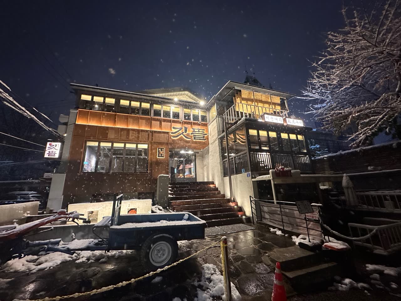눈 내리는 밤의 깃뜰 홍대본점. 목조 2층 건물 창마다 불이 켜져 있고 갈색 벽면에 금빛 깃뜰 간판이 걸려 있다. 돌계단 앞에는 작은 짐차가 세워져 있고 오른쪽 나뭇가지에 눈이 쌓인 홍대 고깃집의 겨울밤 외관